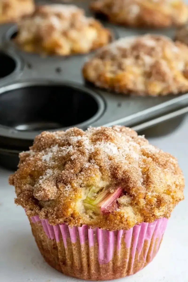Close-up of a Rhubarb Muffin with Greek Yogurt in a pink liner, topped with a cinnamon-sugar crumble and visible rhubarb chunks