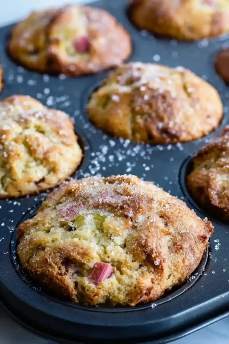 Overhead view of Rhubarb Muffins with Greek Yogurt in a nonstick tin, golden-brown tops sprinkled with sugar and visible rhubarb pieces
