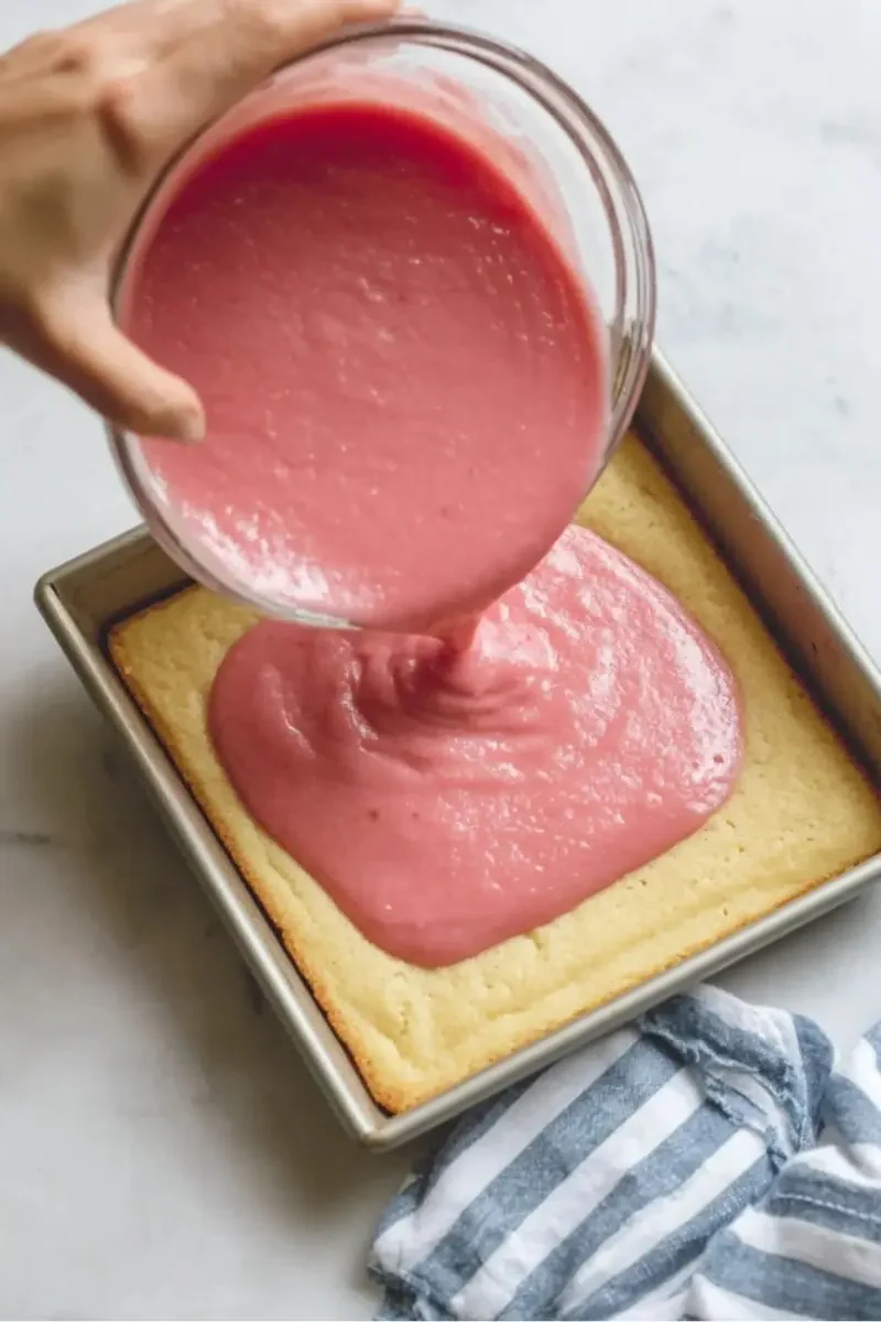 Hand pouring bright pink rhubarb filling over a golden shortbread base in a square baking pan