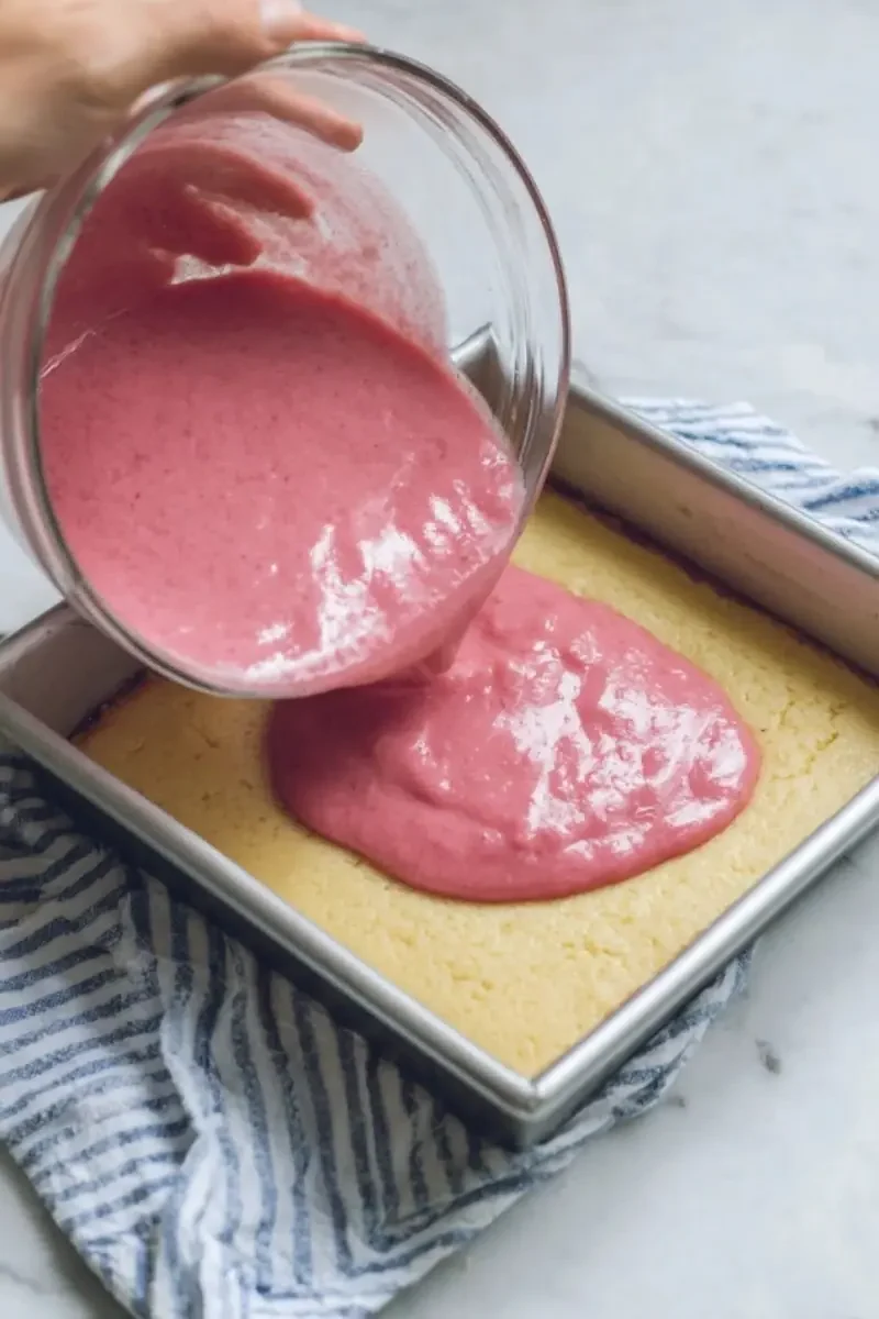 Hand pouring bright pink rhubarb filling over a crisp, golden shortbread base in a square baking pan