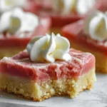 Close-up of square Rhubarb Shortbread Bars with a buttery shortbread base, vibrant pink rhubarb layer, and a swirl of whipped cream on top