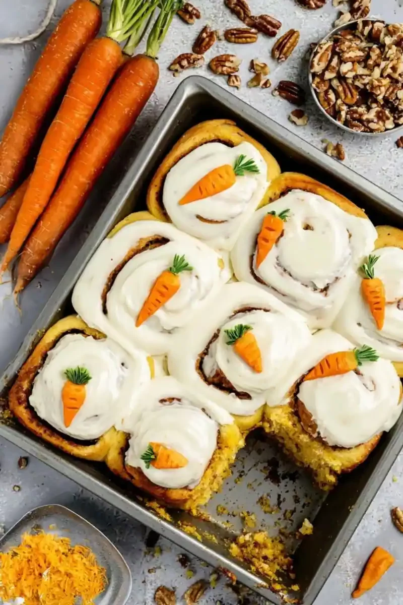 Homemade carrot cake cinnamon rolls in a baking pan topped with cream cheese frosting and fondant carrot decorations